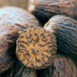 A close up of a pile of nutmeg seeds. One has be cut down the center to reveal the inside of the seed. It is light in color with a darker brown vein-like pattern.