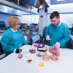 Two scientists standing next to each other at a large table in the kitchen examining different foods.