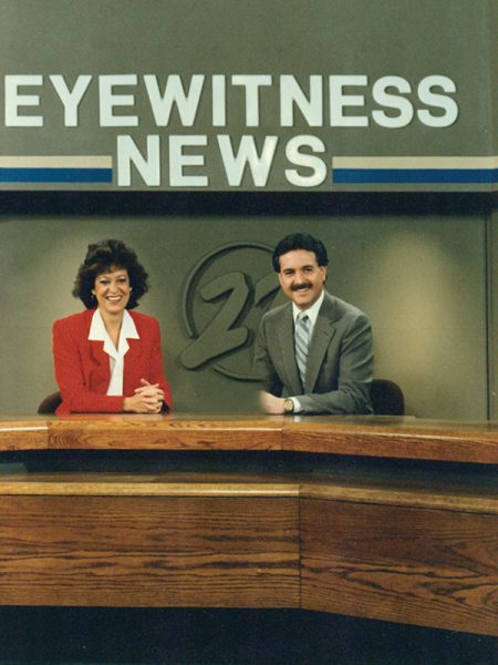 A man and a women sit a desk on the set of a news tv show from decades ago. 