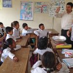 A classroom with lots of young students sitting at their desks and two adults standing at the front of the room.