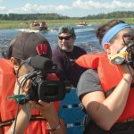 Students on a boat in life jackets using large cameras and recording