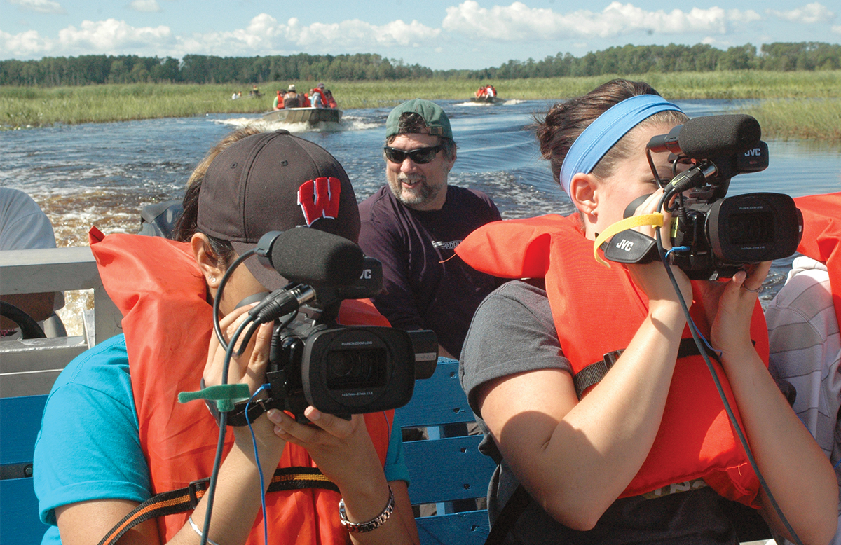 Students on a boat in life jackets using large cameras and recording