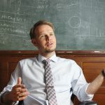 A man siting at a desk in front of a large chalk board with writing on it