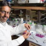 A scientist in a lab coat sitting at a lab table that's covered in small bottles, syringes, and test tubes
