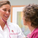 Jacquelynn wearing a lab coat and stethoscope aiding a patient in her office