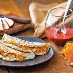 A display of a plate of quesadilla, a glass bowl of oranges, and a cup of a thick Mexican soup
