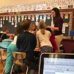 A teacher in a classroom surrounded by a collection of young students as she explains something
