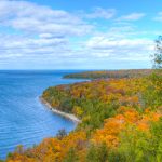 A lake shore. The land is covered in trees that are changing colors with the seasons.