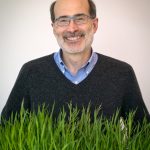 Rick Amasino smiling in front of a plain background. A chunk of grass sits in front of him framing the photo.