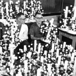 An older black and white photo of two men sitting among large diagrams of molecules