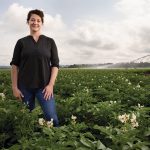 A woman stands, smiling for the camera, in a potato field that stretches far past her into the distance.