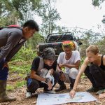 A group of four squatting on the forest floor around a map. One woman points down at the map as the group talks.
