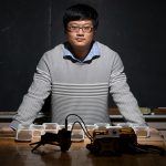 Jingyi Huang stands in his lab with two rows of soil samples in dishes and soil-sensing equipment.