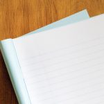 The top left corner of an exam blue book sitting on a wooden desk. The cover of the book is open and folder under the ruled pages.