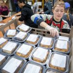 Undergraduate wearing a hair net and rubber gloves places meals packaged in plastic containers on top of a rack.