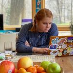 Young woman works on her laptop computer at a table in a bright, naturally lit kitchen.