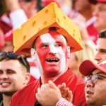 A UW Badger fan with face painted red and white and wearing a cheesehead stands in a crowd.