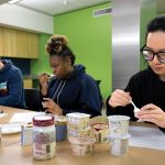 Three students sitting at a table in a classroom tasting types of yogurt.