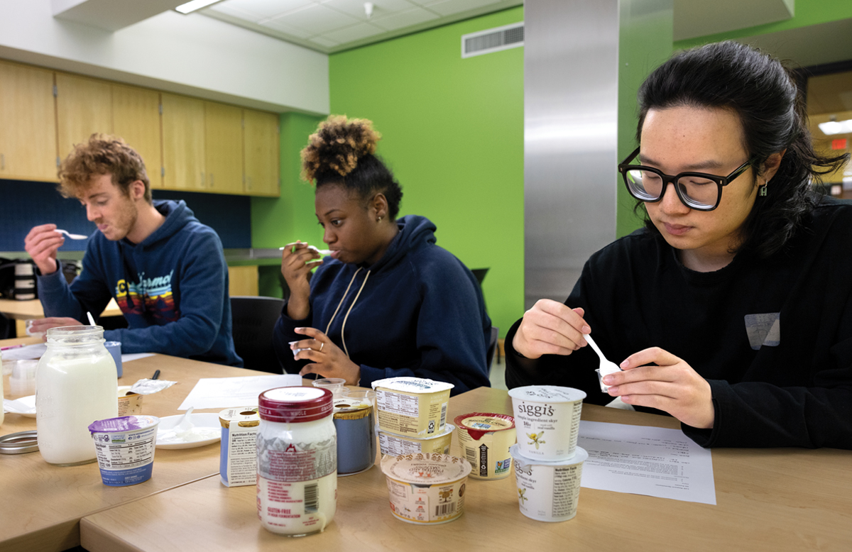 Three students sitting at a table in a classroom tasting types of yogurt.