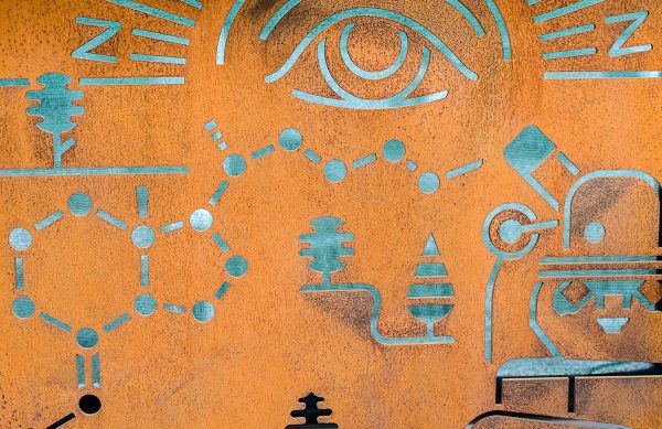 A rusty metal art piece with different shaped cut outs. There are chemical cut outs, scientific equipment, and an eye.
