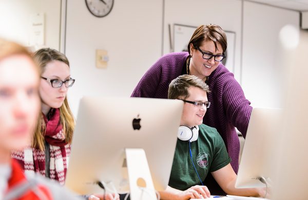 A woman stands behind a seated male student and points at the computer screen they are both viewing.