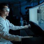 A student in a lab coat and goggles at a desk working on a desktop computer.
