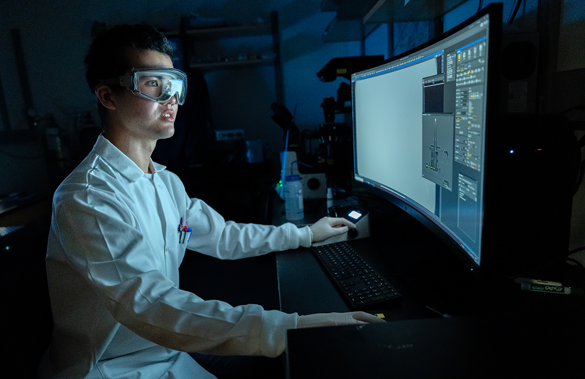 A student in a lab coat and goggles at a desk working on a desktop computer.