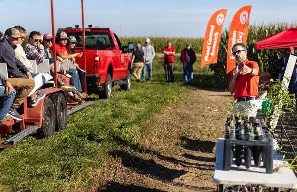 A trolly of people sit and listen to a man. The man gestures to a table of plant samples and talks in a microphone to the trolly of people.