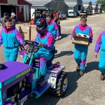 Five members of the pull team dressed in matching colorful windbreakers. They carry equipment and drive their tractor through a lot.