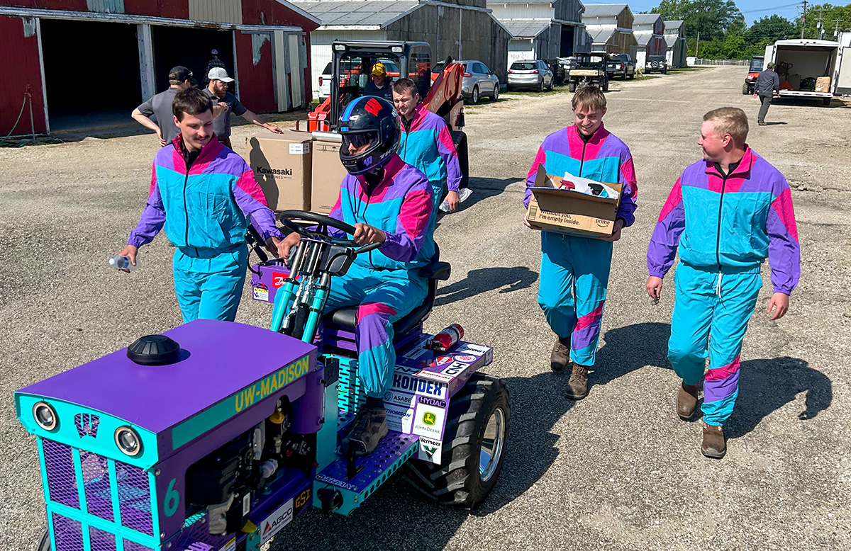 Five members of the pull team dressed in matching colorful windbreakers. They carry equipment and drive their tractor through a lot.