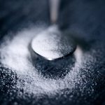 The bowl of a silver-colored spoon, filled with white granules, rests upon a shallow pile of more white granules.