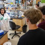 A woman in a a white lab coat stands near a bench and shelves with glassware in a lab as she speaks to a small group.