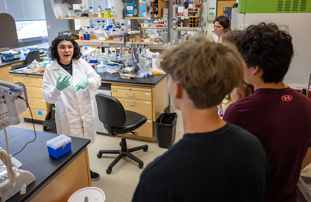 A woman in a a white lab coat stands near a bench and shelves with glassware in a lab as she speaks to a small group.