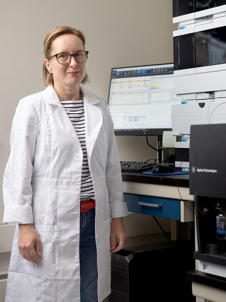 A professor in a lab coat stands in front of a computer and machinery in a lab
