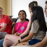 A panel of five students sit in a line. A girl holds a microphone and speaks. The other students in the line look at her and smile as she speaks.