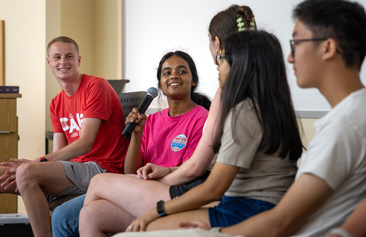 A panel of five students sit in a line. A girl holds a microphone and speaks. The other students in the line look at her and smile as she speaks.