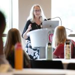 Dean Glenda Gillaspy Stands at a podium in the front of a room. She speaks into a microphone while student sit at tables listening to her.