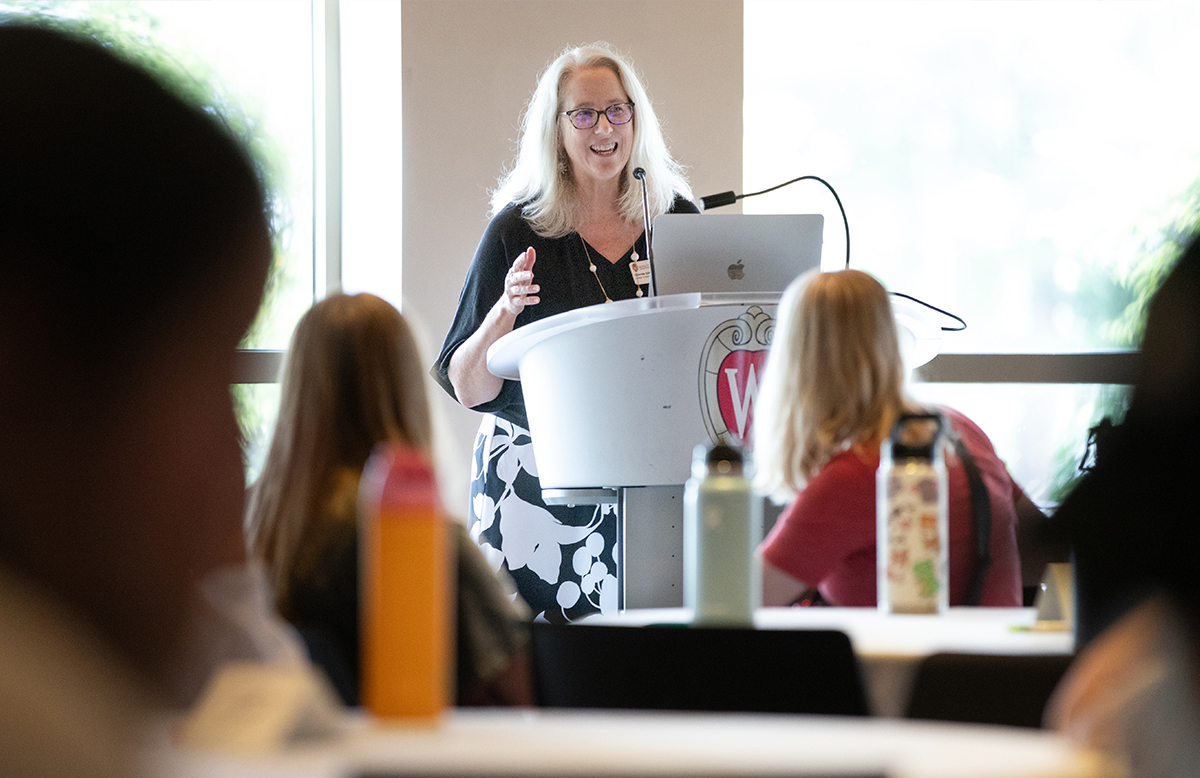 Dean Glenda Gillaspy Stands at a podium in the front of a room. She speaks into a microphone while student sit at tables listening to her.