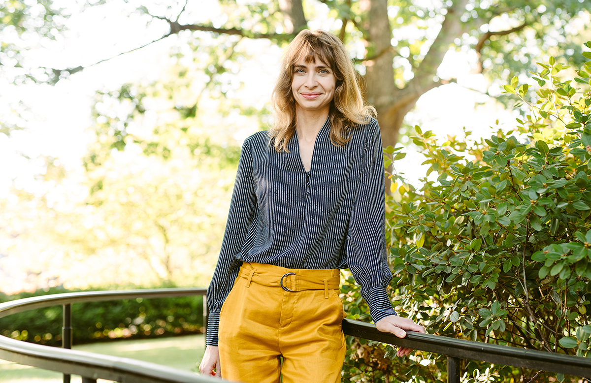 A woman smiles while leaning up against a metal railing along a path in a wooded garden setting.