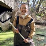 A woman wears waders in a bog. She holds up a net and smiles.