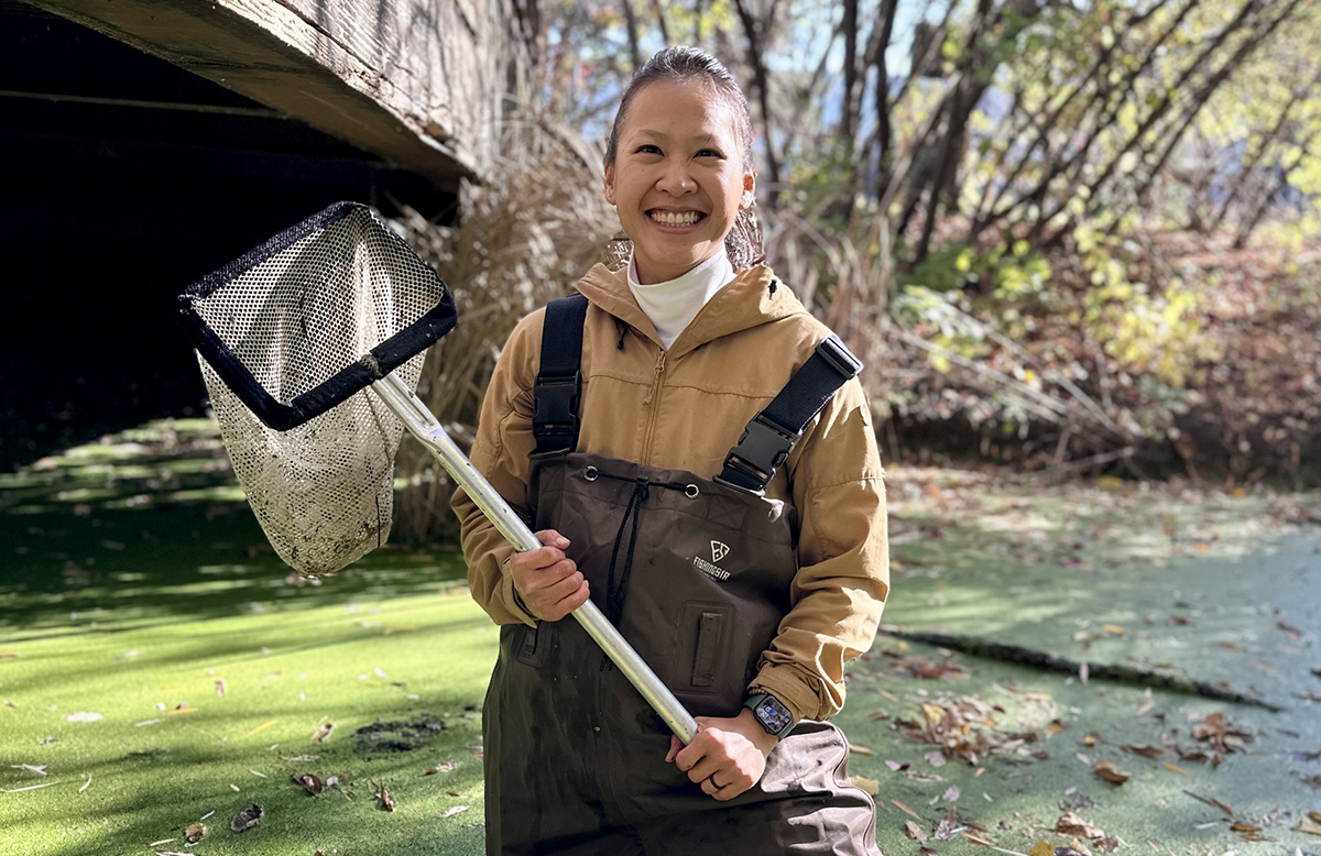 A woman wears waders in a bog. She holds up a net and smiles.