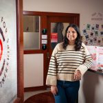 A view of the entrance to the Center for Patient Partnerships. On the door there is a logo. Inside the office, a student employee leans up against their front desk.