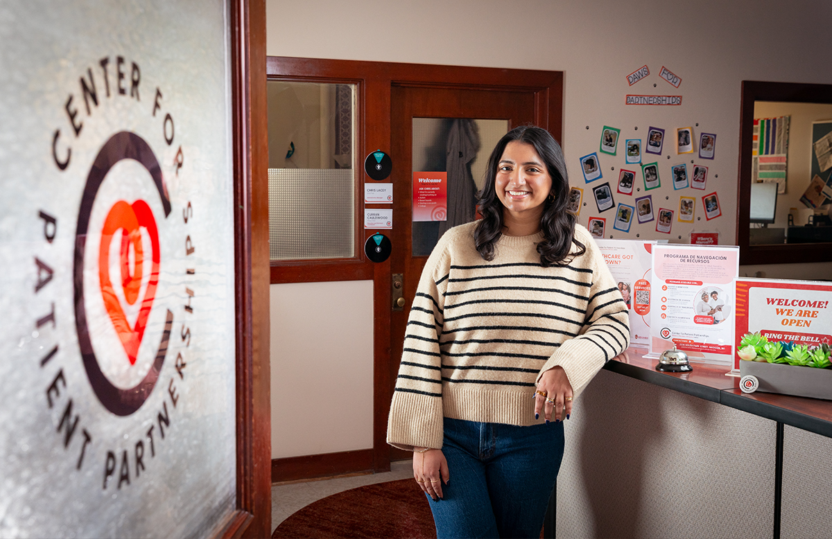 A view of the entrance to the Center for Patient Partnerships. On the door there is a logo. Inside the office, a student employee leans up against their front desk.
