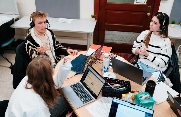 Three student employees sit together around a table. They have paper, notebooks, and laptops. Two of the students wear headphones as they work. 