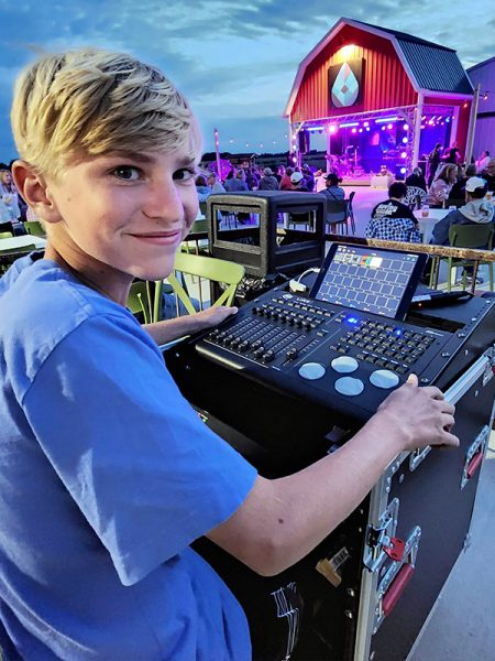 A young boy sitting behind a sound mixing board. In front of him is a stage with a band performing. 