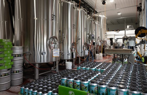 A row of kegs. In front of them are stacks of hundreds of cans of beer. 
