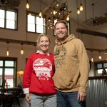 a man and woman standing next to each other, smiling. They are standing inside their business. There is a counter and tables behind them.