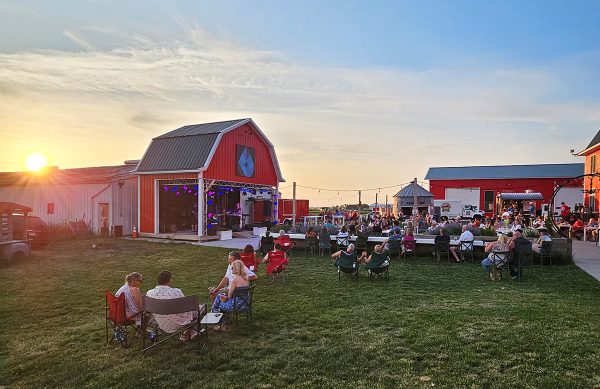A stage, designed like a barn. A band plays to many groups of people in the outdoor space behind other barns. 