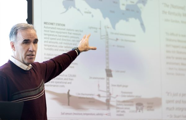 A man stands in front of a screen. One the screen there is a projection of a presentation. He holds up an arm to gesture to the presentation.
