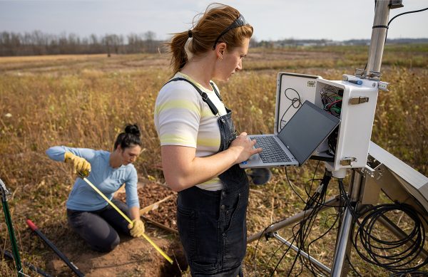 A scienctist stands at an electrical box holding a laptop and working. This is in a field of farmland. Behind her is another person on her hands and knees with a measuring tape.